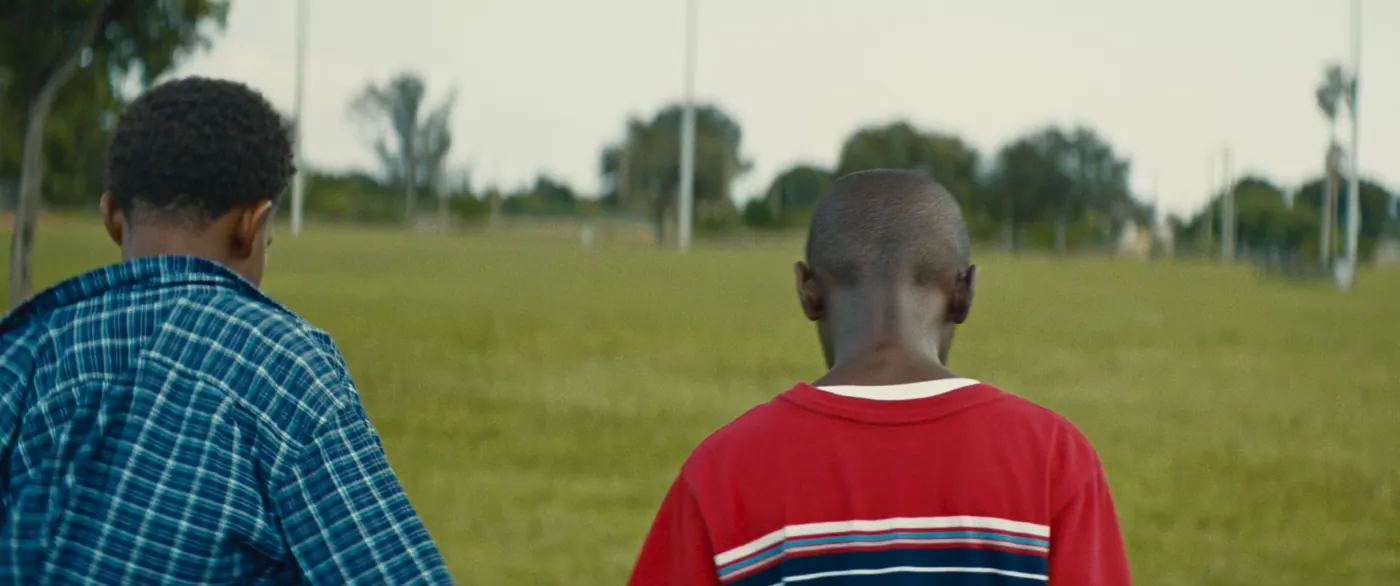 Moonlight (2016), Barry Jenkins — plan rapproché, jour, extérieur, deux personnages : terrain de sport, tee-shirt rouge, chemise à carreaux bleue, palmier, marcher