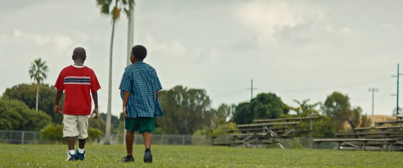 Moonlight (2016), Barry Jenkins — plan large, jour, extérieur, deux personnages : terrain de sport, tee-shirt rouge, short vert, palmier, gradin, grillage, poteau électrique, baskets