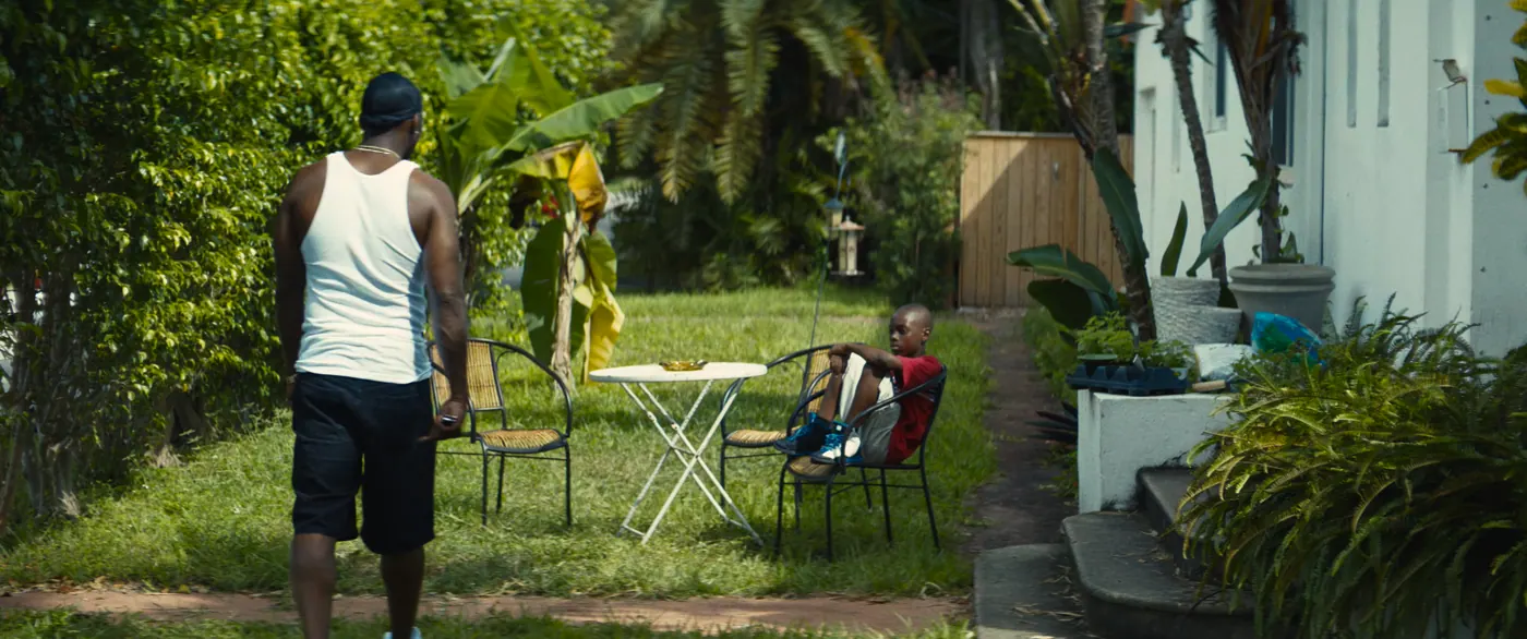 Moonlight (2016), Barry Jenkins — plan large, jour, extérieur, deux personnages : jardin, débardeur blanc, short noir, chaise de jardin, table de jardin, bananier, palmier, plante en pot