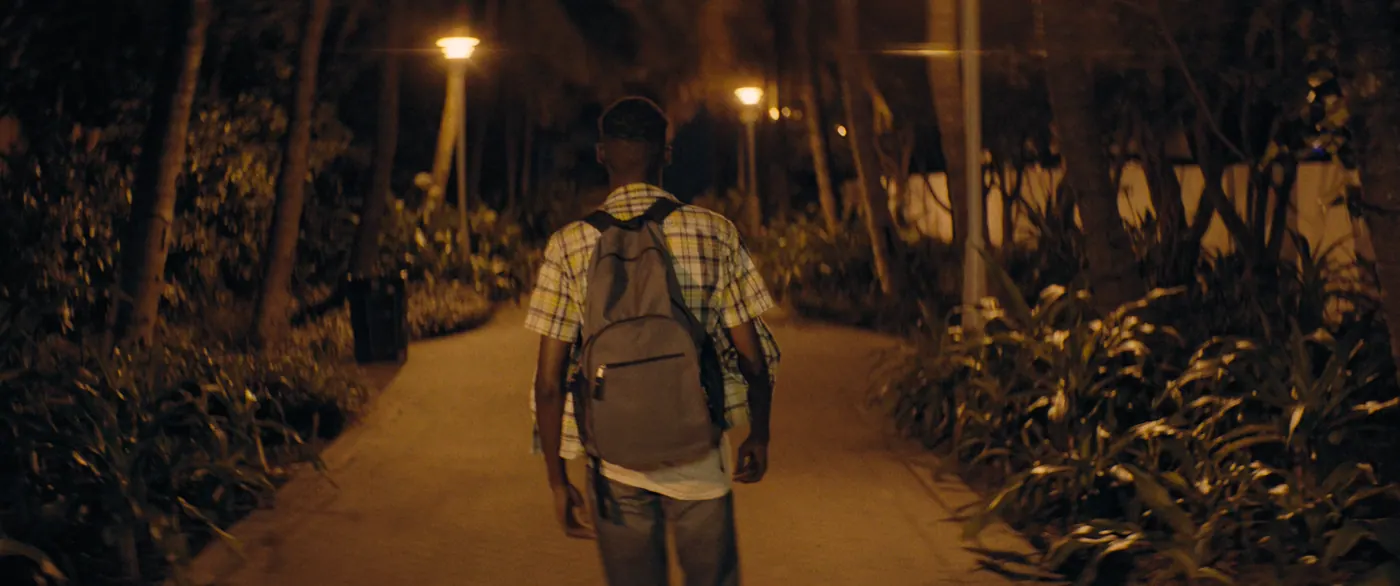 Moonlight (2016), Barry Jenkins — plan moyen, nuit, extérieur, un personnage, lumière chaude : parc, lampadaire, sac à dos, poubelle, palmier, chemise à carreaux, marcher