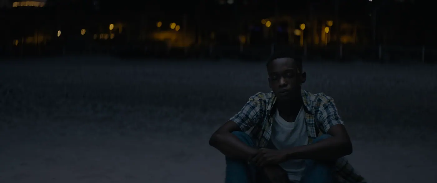 Moonlight (2016), Barry Jenkins — plan moyen, nuit, extérieur, un personnage, lumière froide : plage, tee-shirt blanc, chemise à carreaux, jean