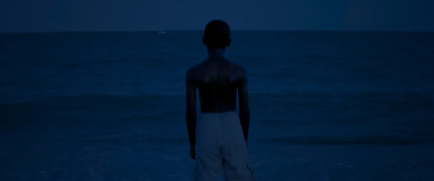 Moonlight (2016), Barry Jenkins — plan moyen, nuit, extérieur, un personnage, lumière froide : plage, mer, torse nu, short