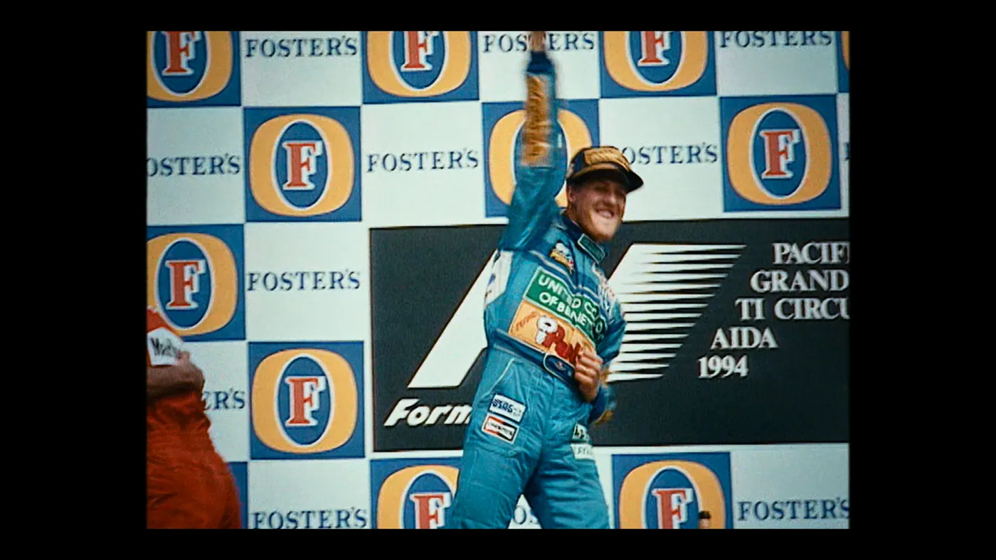 Schumacher ’94 — The Birth of a Legend (2026), Christin Freitag — plan moyen, jour, extérieur, deux personnages : podium, circuit automobile, combinaison bleue, panneau publicitaire