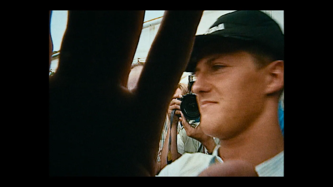 Schumacher ’94 — The Birth of a Legend (2026), Christin Freitag — gros plan, jour, extérieur, quatre personnages : casquette noire, appareil photo, prendre une photo
