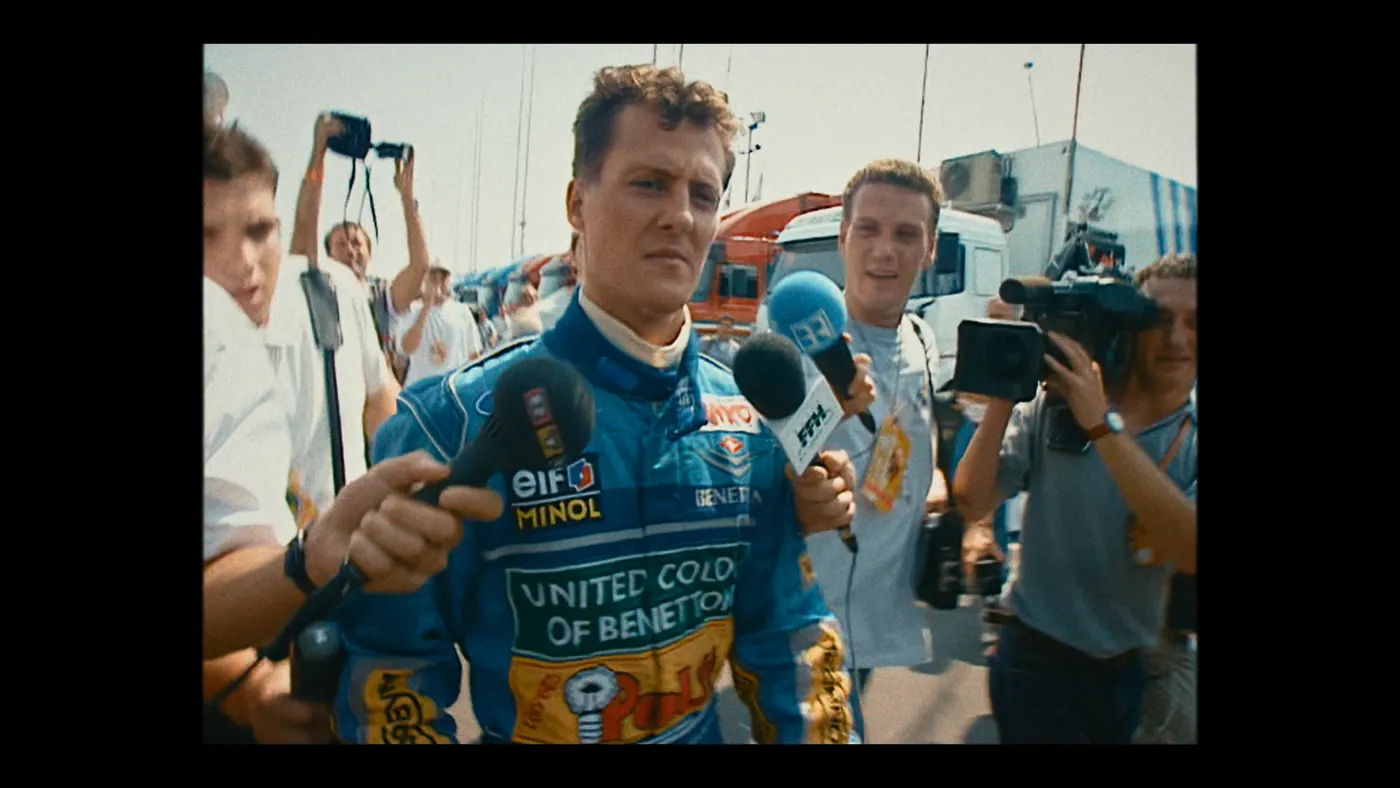 Schumacher ’94 — The Birth of a Legend (2026), Christin Freitag — plan rapproché, jour, extérieur, foule : paddock, circuit automobile, combinaison bleue, micro, caméra, appareil photo