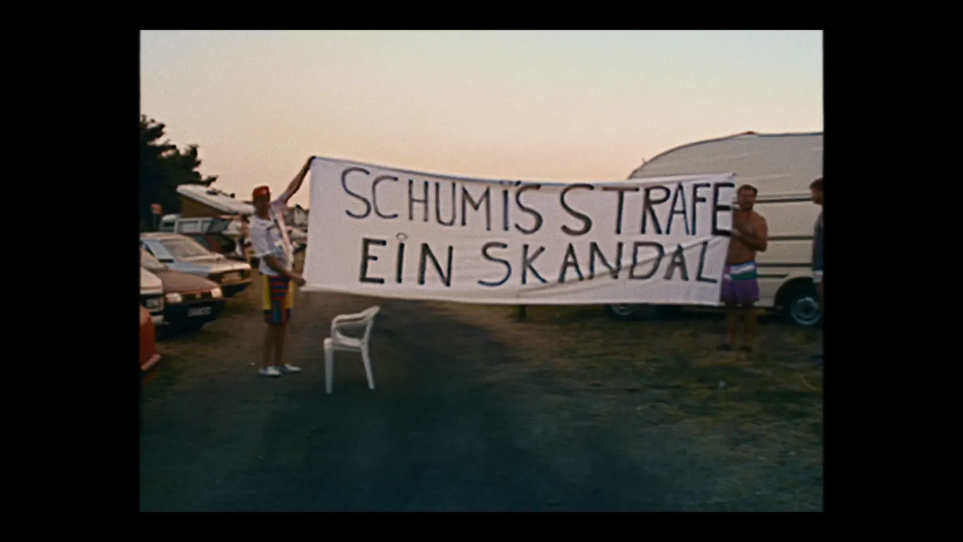 Schumacher ’94 — The Birth of a Legend (2026), Christin Freitag — plan large, aube ou crépuscule, extérieur, trois personnages : camping, banderole, caravane, voiture, chaise monobloc, arbre