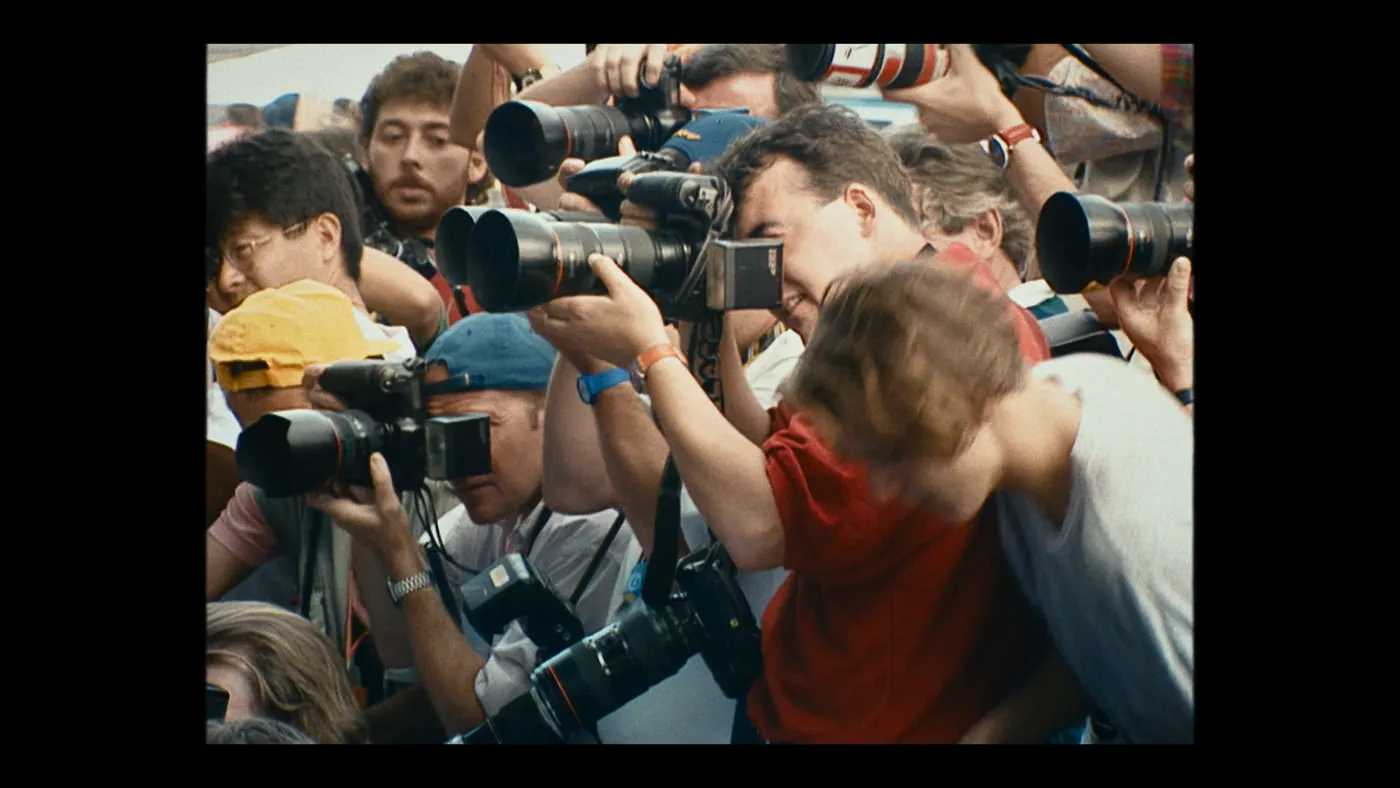 Schumacher ’94 — The Birth of a Legend (2026), Christin Freitag — plan rapproché, jour, extérieur, foule : casquette jaune, appareil photo, téléobjectif, montre, lunettes, prendre une photo