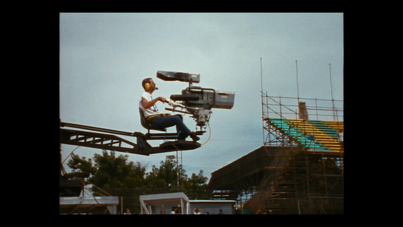 Schumacher ’94 — The Birth of a Legend (2026), Christin Freitag — plan large, jour, extérieur, foule : tribune, caméra, grue de caméra, échafaudage, arbre, casque audio, tee-shirt, filmer