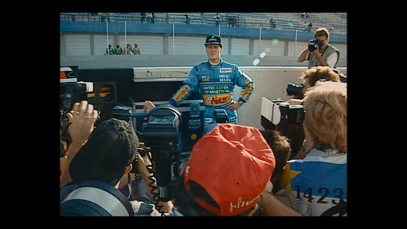 Schumacher ’94 — The Birth of a Legend (2026), Christin Freitag — plan moyen, jour, extérieur, foule, soleil direct : circuit automobile, tribune, casquette rouge, combinaison bleue