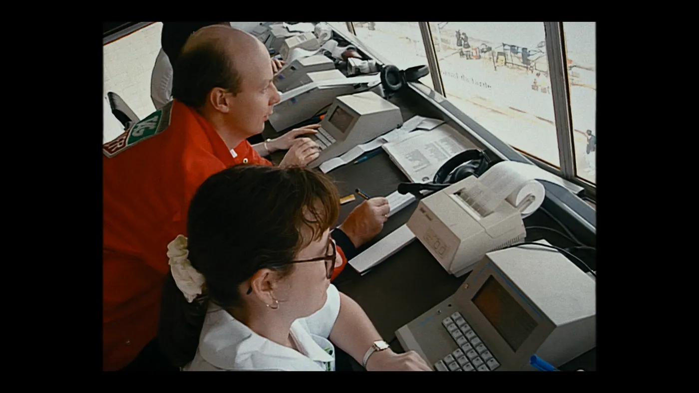 Schumacher ’94 — The Birth of a Legend (2026), Christin Freitag — plan rapproché, jour, intérieur, trois personnages, lumière de fenêtre : salle de contrôle, veste rouge, imprimante, clavier