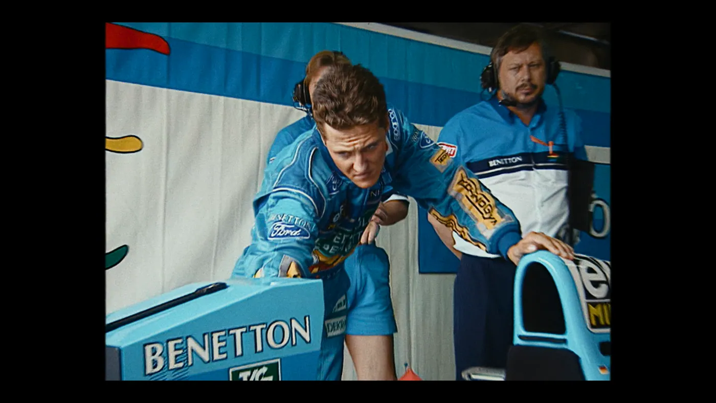 Schumacher ’94 — The Birth of a Legend (2026), Christin Freitag — plan moyen, intérieur, trois personnages : garage, circuit automobile, combinaison bleue, voiture de course, formule 1