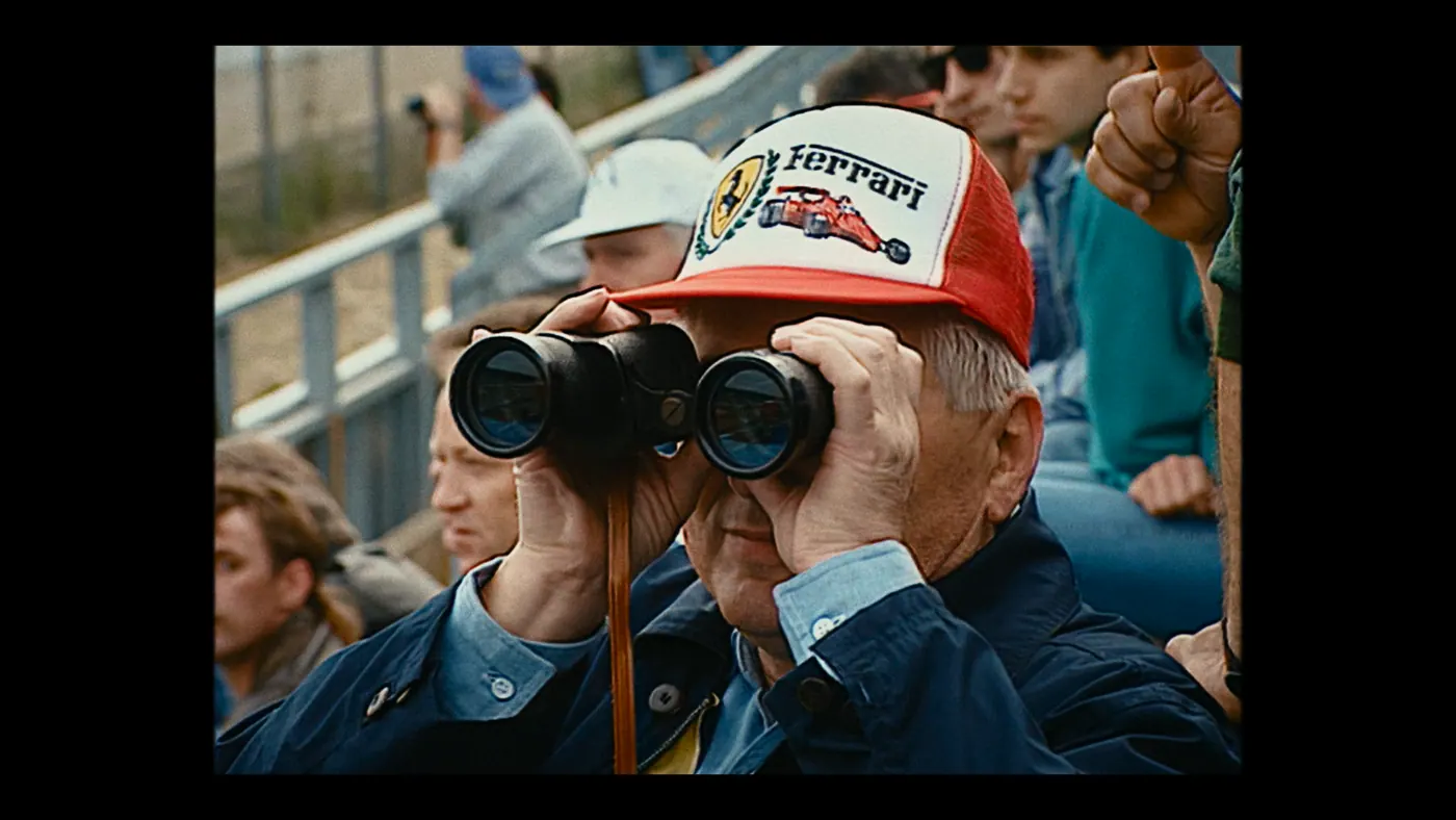 Schumacher ’94 — The Birth of a Legend (2026), Christin Freitag — plan rapproché, jour, extérieur, foule : tribune, jumelles, casquette, blouson, regarder aux jumelles