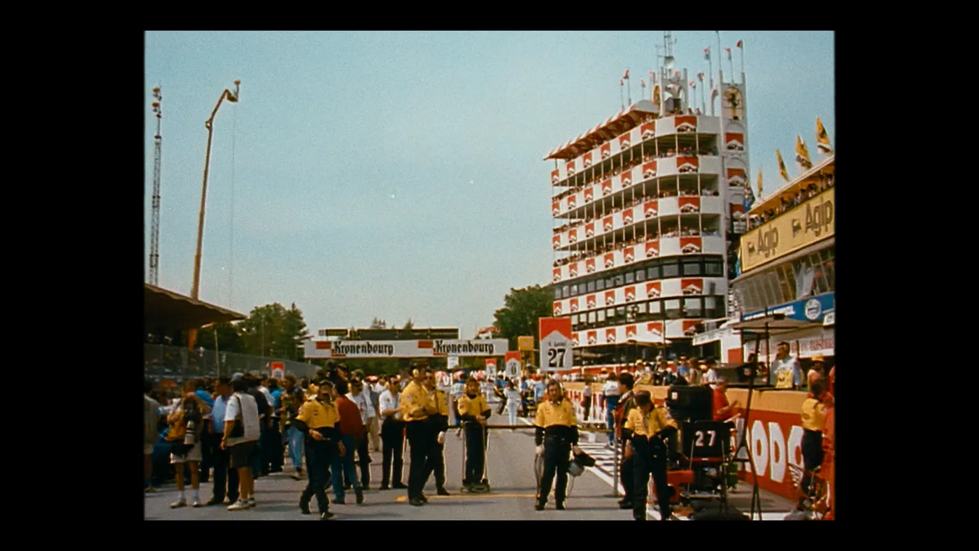 Schumacher ’94 — The Birth of a Legend (2026), Christin Freitag — plan d’ensemble, jour, extérieur, foule, soleil direct : circuit automobile, grille de départ, uniforme jaune, drapeau