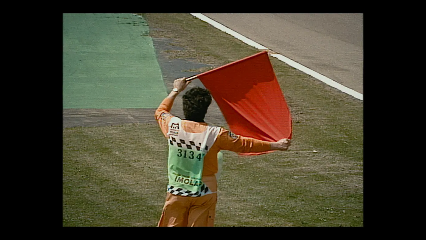 Schumacher ’94 — The Birth of a Legend (2026), Christin Freitag — plan moyen, jour, extérieur, un personnage, soleil direct : circuit automobile, combinaison orange, drapeau rouge, dossard