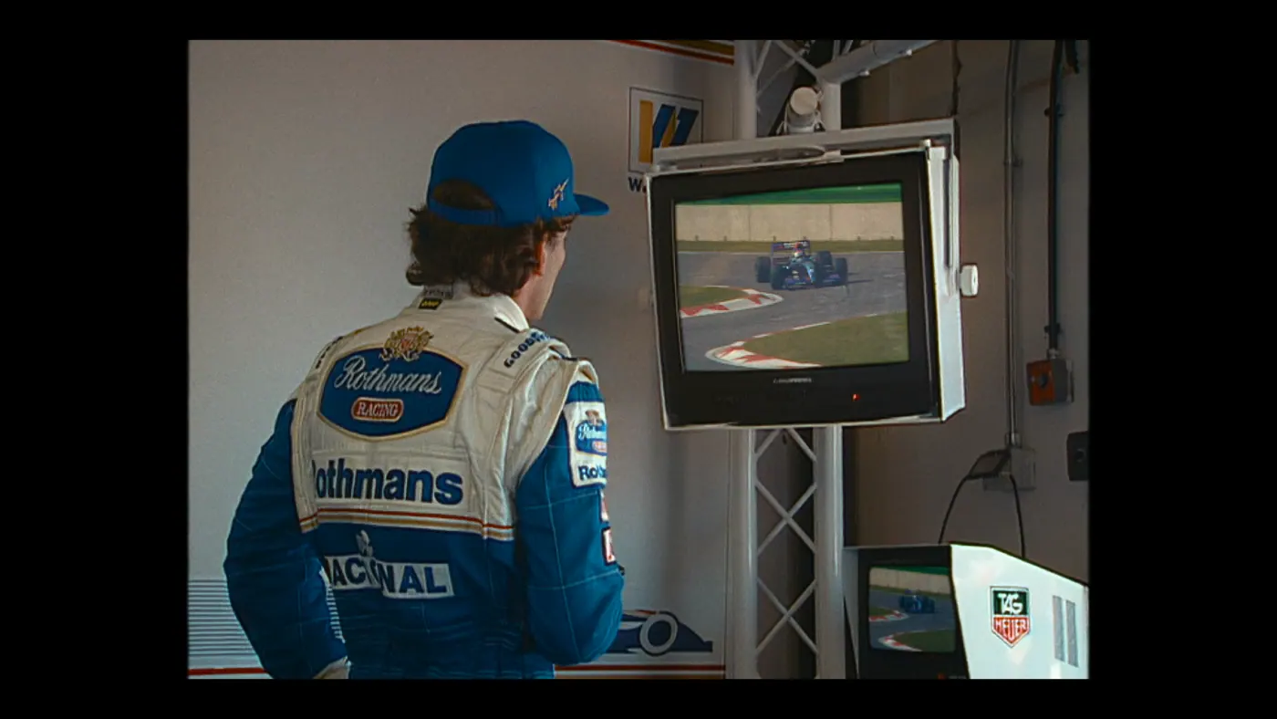 Schumacher ’94 — The Birth of a Legend (2026), Christin Freitag — plan moyen, intérieur, un personnage : garage, circuit automobile, casquette bleue, téléviseur, voiture de course, formule 1