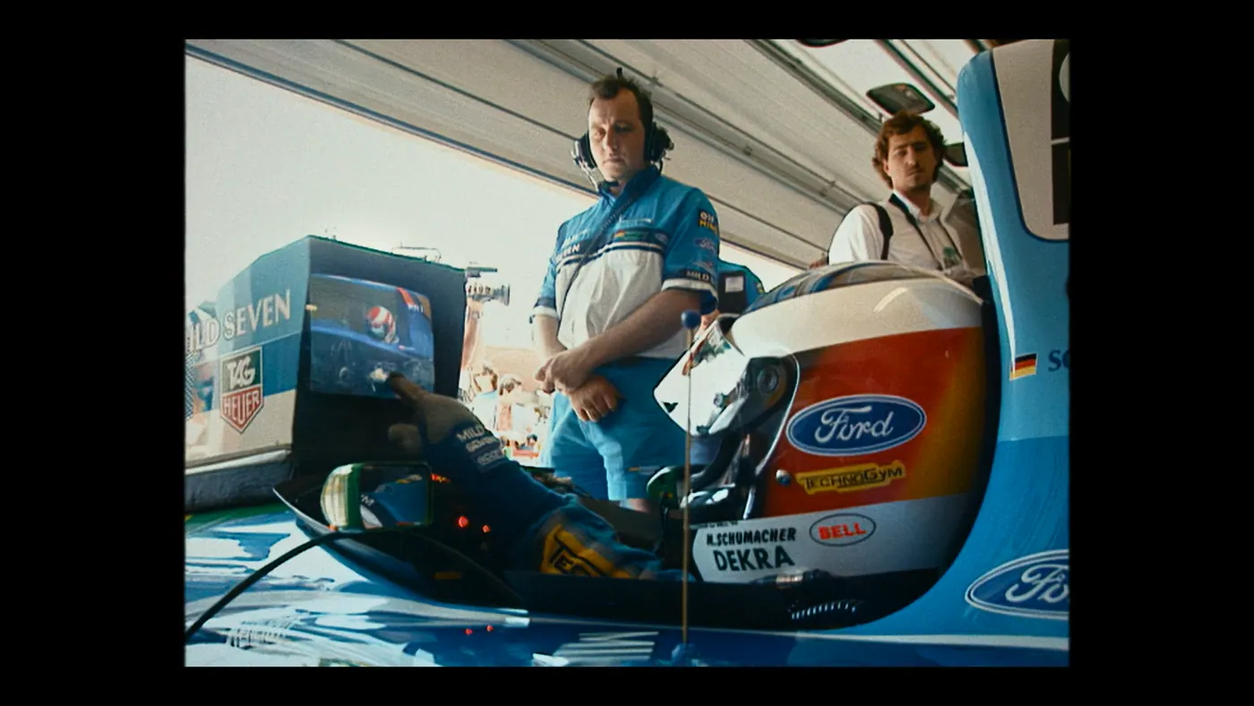 Schumacher ’94 — The Birth of a Legend (2026), Christin Freitag — plan moyen, jour, intérieur, trois personnages : garage, voiture (habitacle), voiture de course, formule 1, téléviseur