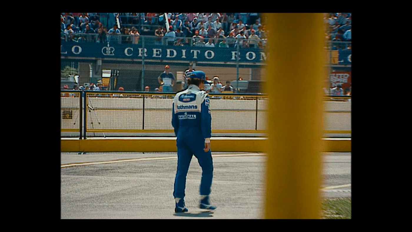 Schumacher ’94 — The Birth of a Legend (2026), Christin Freitag — plan large, jour, extérieur, foule, soleil direct : circuit automobile, tribune, combinaison bleue, grillage