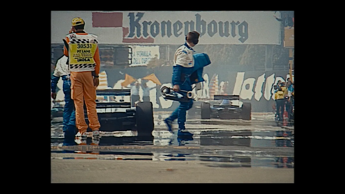 Schumacher ’94 — The Birth of a Legend (2026), Christin Freitag — plan large, jour, extérieur, foule : circuit automobile, combinaison orange, casquette jaune, voiture de course, formule 1