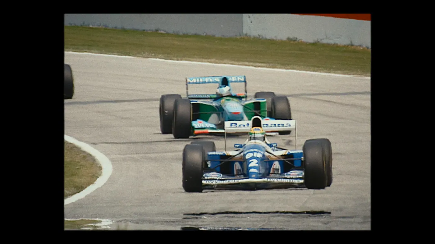 Schumacher ’94 — The Birth of a Legend (2026), Christin Freitag — plan large, jour, extérieur, deux personnages, soleil direct : circuit automobile, voiture de course, formule 1, casque