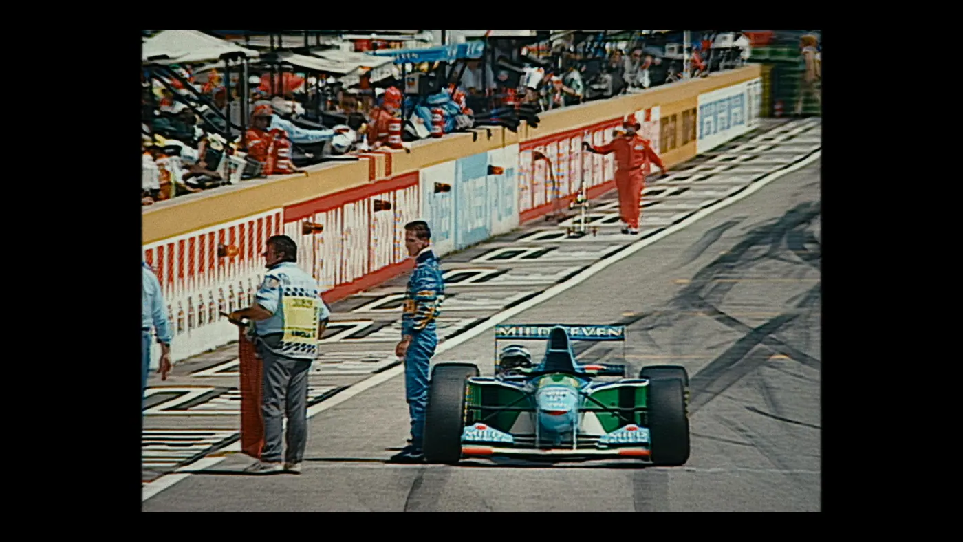 Schumacher ’94 — The Birth of a Legend (2026), Christin Freitag — plan large, jour, extérieur, foule, soleil direct : circuit automobile, combinaison rouge, combinaison bleue