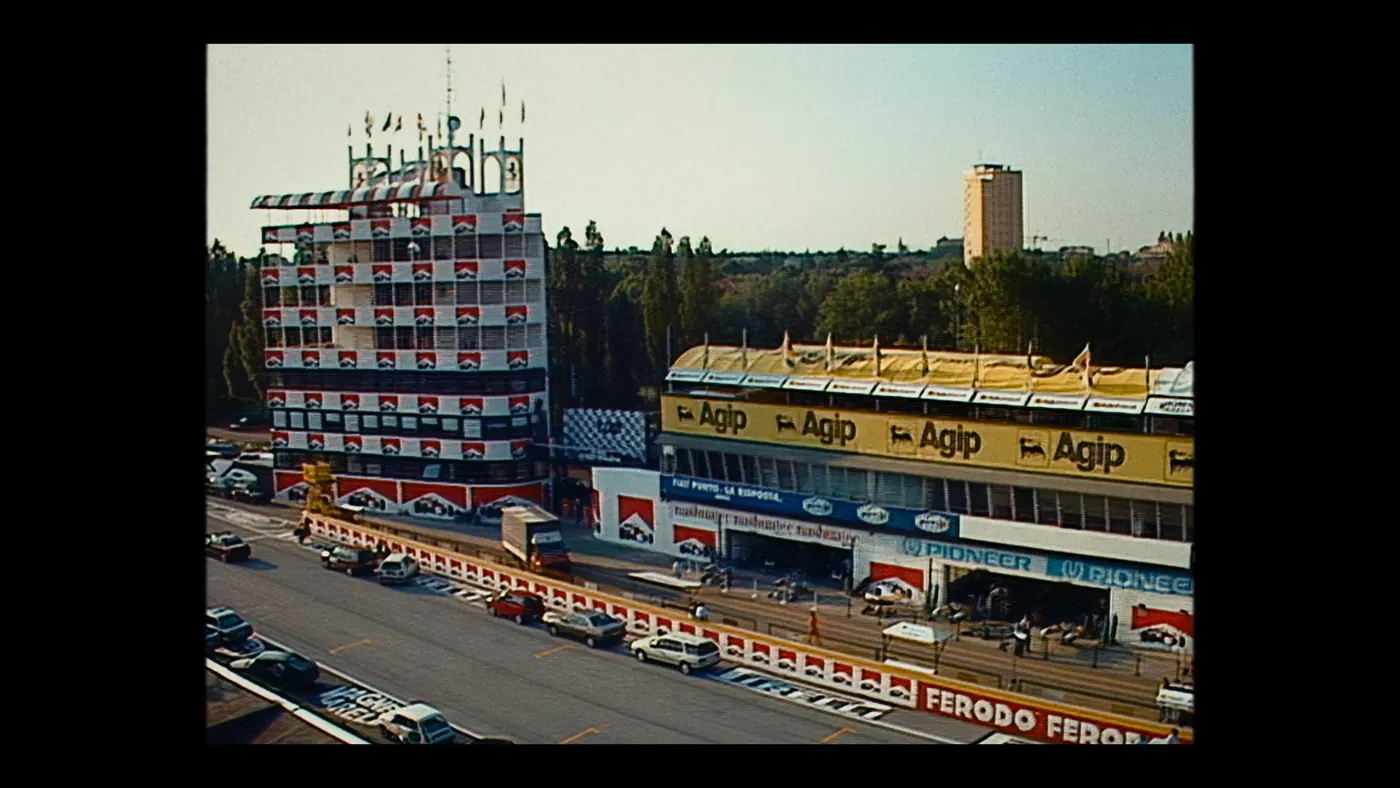 Schumacher ’94 — The Birth of a Legend (2026), Christin Freitag — plan d’ensemble, jour, extérieur, foule : circuit automobile, tour, drapeau, panneau publicitaire, voiture, camion