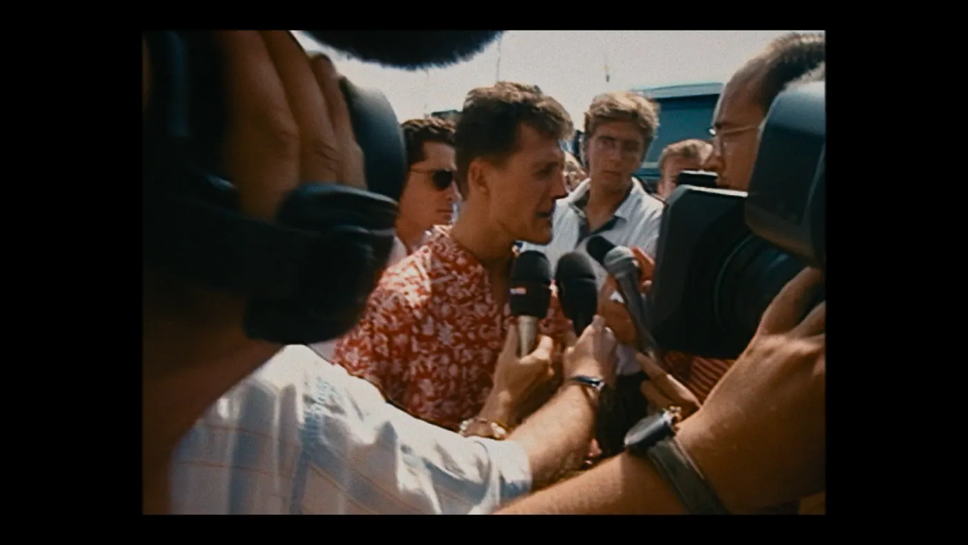Schumacher ’94 — The Birth of a Legend (2026), Christin Freitag — plan rapproché, jour, extérieur, foule : chemise rouge, micro, caméra, montre, chemise hawaïenne, lunettes de soleil