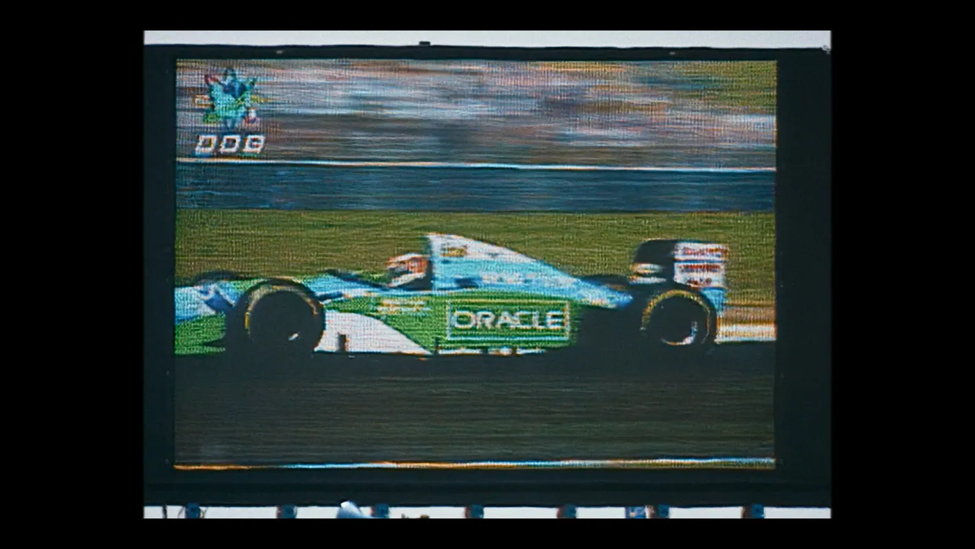 Schumacher ’94 — The Birth of a Legend (2026), Christin Freitag — insert, jour, extérieur, un personnage : circuit automobile, écran géant, voiture de course, formule 1, casque, conduire