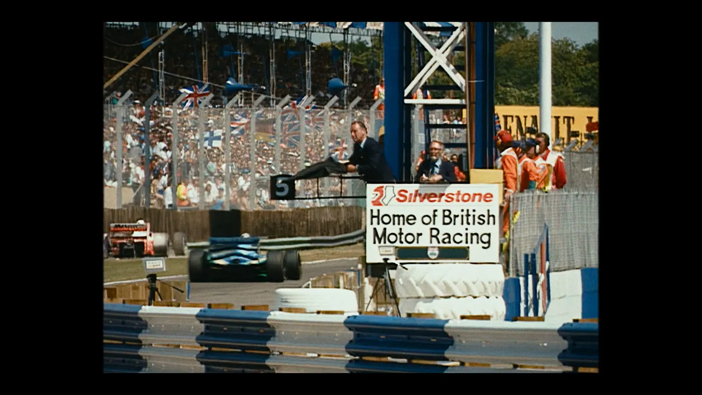 Schumacher ’94 — The Birth of a Legend (2026), Christin Freitag — plan large, jour, extérieur, foule, soleil direct : circuit automobile, combinaison orange, voiture de course, formule 1