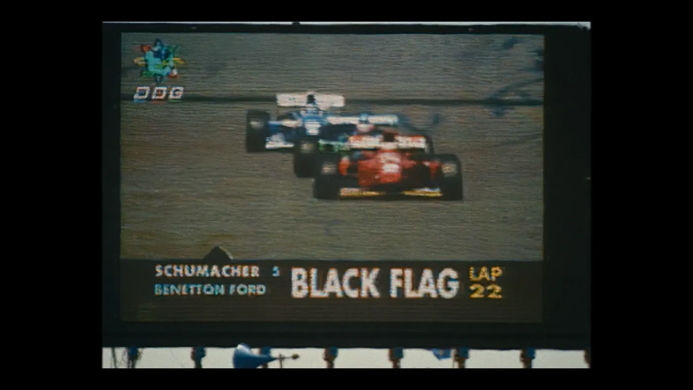 Schumacher ’94 — The Birth of a Legend (2026), Christin Freitag — insert, jour, extérieur, deux personnages : circuit automobile, écran géant, voiture de course, formule 1, casque, conduire