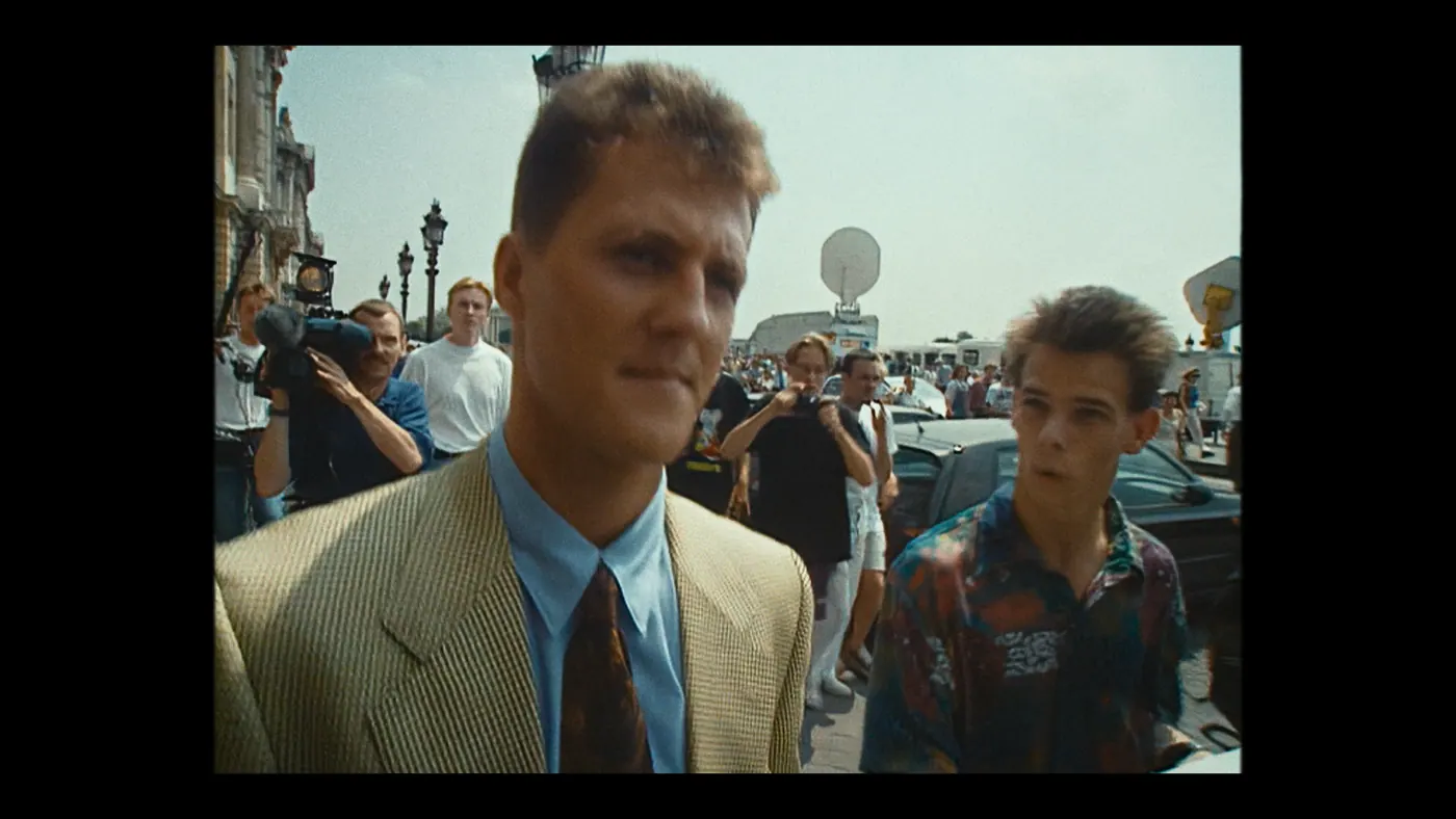 Schumacher ’94 — The Birth of a Legend (2026), Christin Freitag — plan rapproché, jour, extérieur, foule : rue, ville, veste beige, chemise bleue, caméra, appareil photo, antenne parabolique