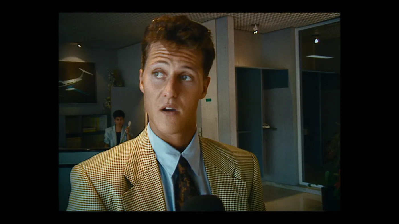 Schumacher ’94 — The Birth of a Legend (2026), Christin Freitag — plan rapproché, intérieur, deux personnages : bureau, veste jaune, chemise bleue, micro, téléphone fixe, avion, tableau