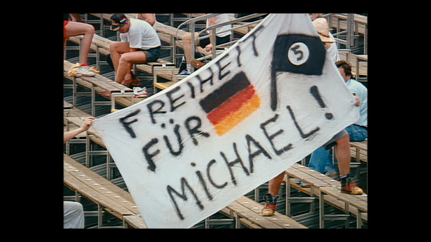 Schumacher ’94 — The Birth of a Legend (2026), Christin Freitag — plan moyen, jour, extérieur, foule : stade, banderole, drapeau allemand, drapeau noir, gradin, casquette, tee-shirt