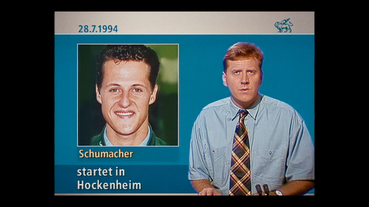 Schumacher ’94 — The Birth of a Legend (2026), Christin Freitag — plan rapproché, intérieur, deux personnages : studio de télévision, chemise bleue, portrait, micro, montre, logo
