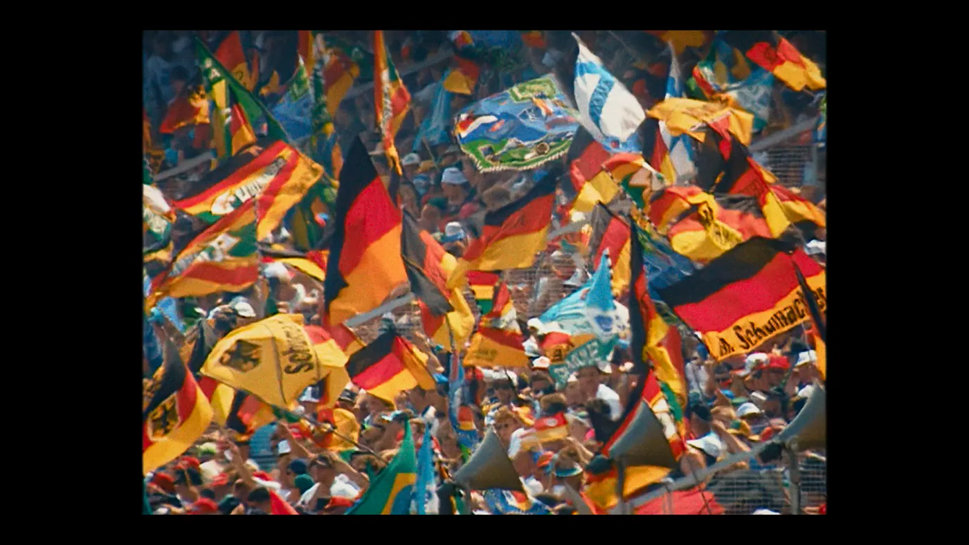 Schumacher ’94 — The Birth of a Legend (2026), Christin Freitag — plan d’ensemble, jour, extérieur, foule : stade, drapeau allemand, drapeau, aigle, haut-parleur, grillage, casquette