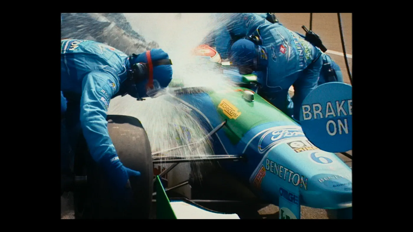 Schumacher ’94 — The Birth of a Legend (2026), Christin Freitag — plan rapproché, jour, extérieur, trois personnages, soleil direct : circuit automobile, combinaison bleue, voiture de course