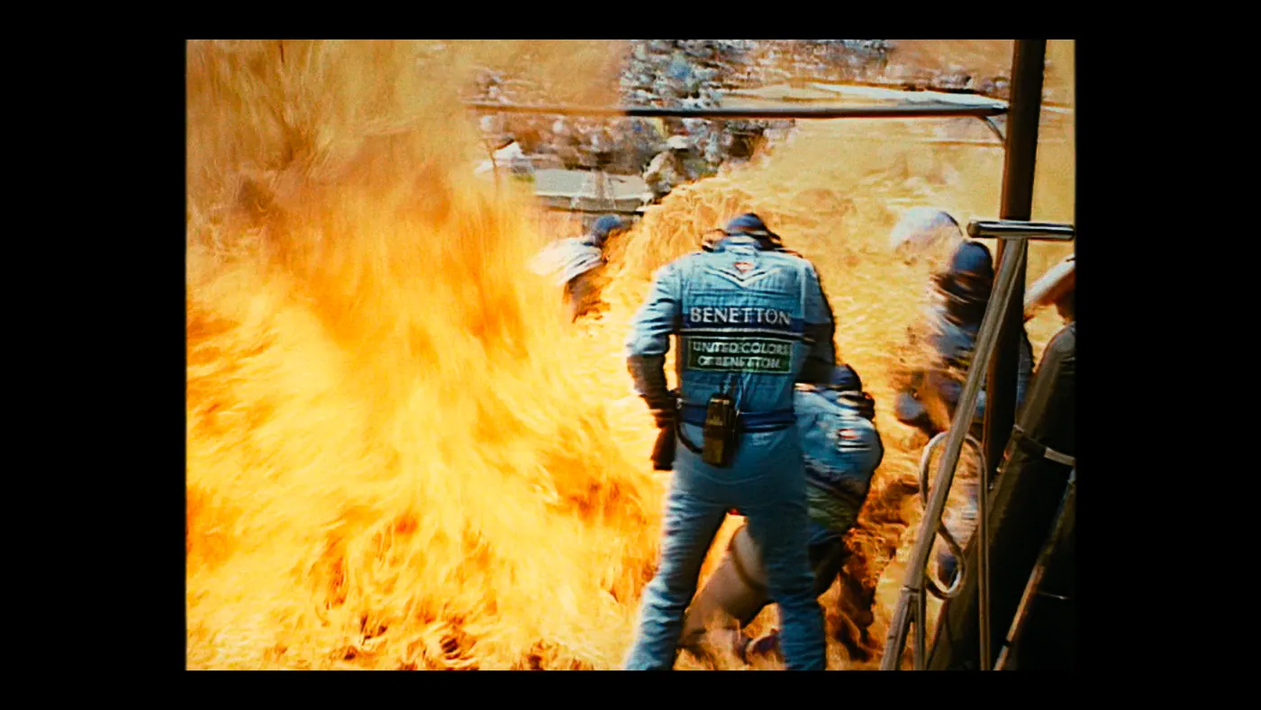 Schumacher ’94 — The Birth of a Legend (2026), Christin Freitag — plan moyen, jour, extérieur, quatre personnages, feu : circuit automobile, combinaison bleue, boule de feu, flamme, casque