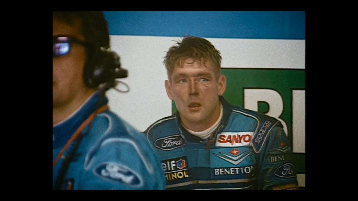 Schumacher ’94 — The Birth of a Legend (2026), Christin Freitag — plan rapproché, jour, deux personnages : combinaison bleue, micro-casque, combinaison de pilote, lunettes