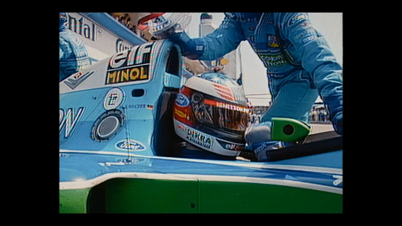Schumacher ’94 — The Birth of a Legend (2026), Christin Freitag — plan rapproché, jour, extérieur, trois personnages, soleil direct : circuit automobile, combinaison bleue, voiture de course