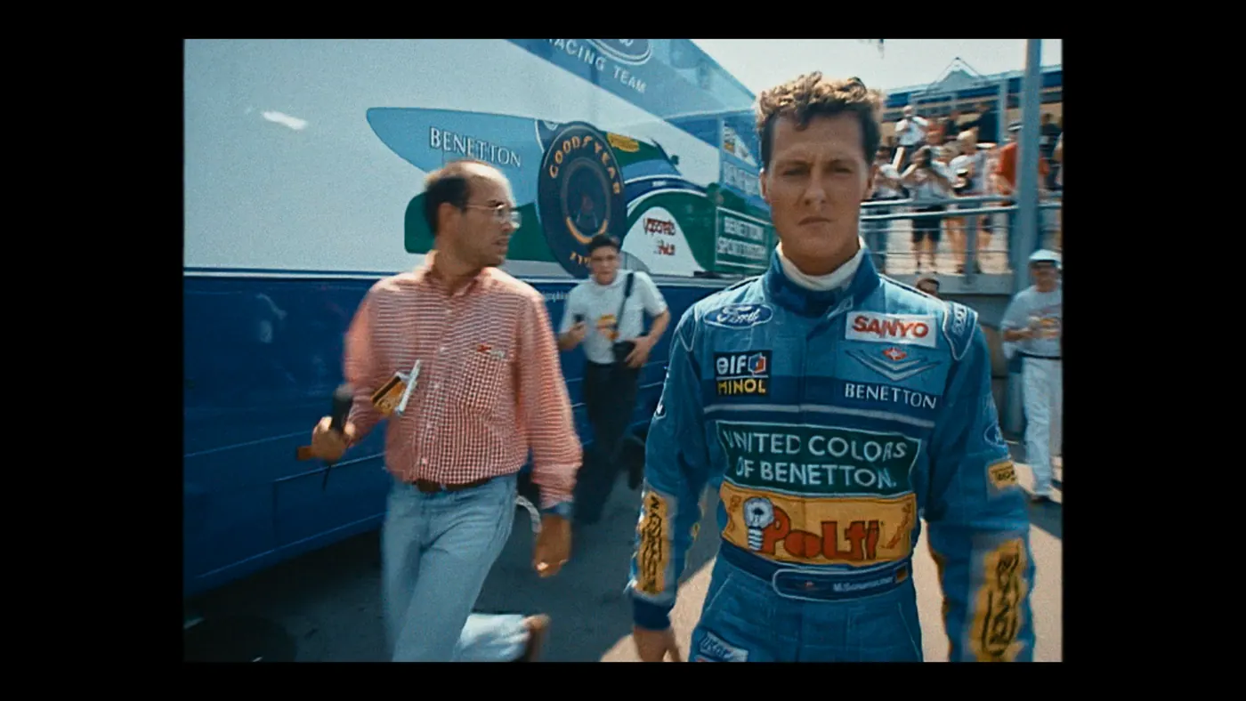 Schumacher ’94 — The Birth of a Legend (2026), Christin Freitag — plan moyen, jour, extérieur, foule, soleil direct : circuit automobile, combinaison bleue, chemise à carreaux rouge, camion