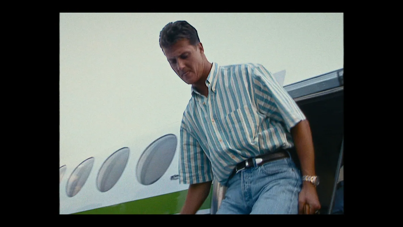 Schumacher ’94 — The Birth of a Legend (2026), Christin Freitag — plan moyen, jour, extérieur, un personnage : avion, hublot, montre, chemise rayée, jean, voyager