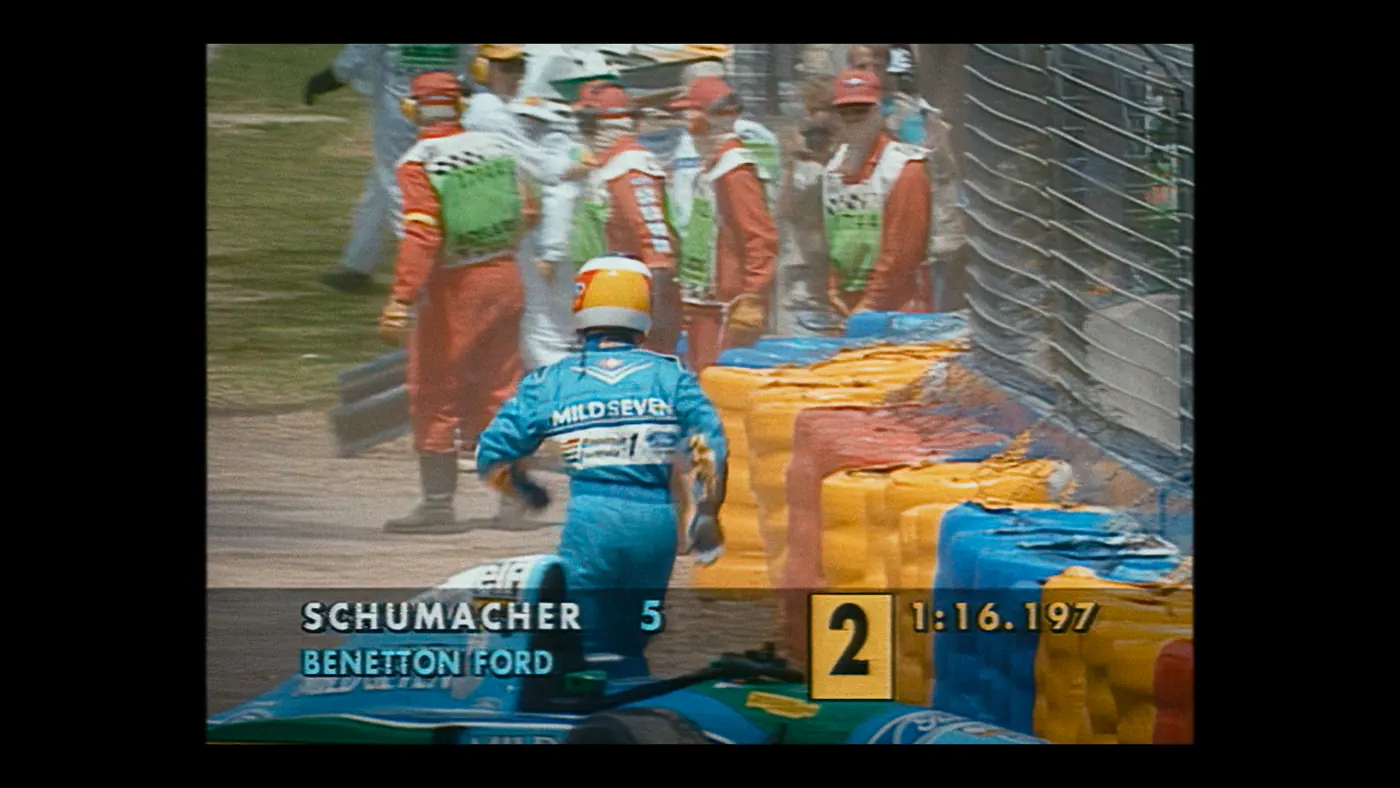 Schumacher ’94 — The Birth of a Legend (2026), Christin Freitag — plan large, jour, extérieur, foule, soleil direct : circuit automobile, combinaison orange, combinaison bleue