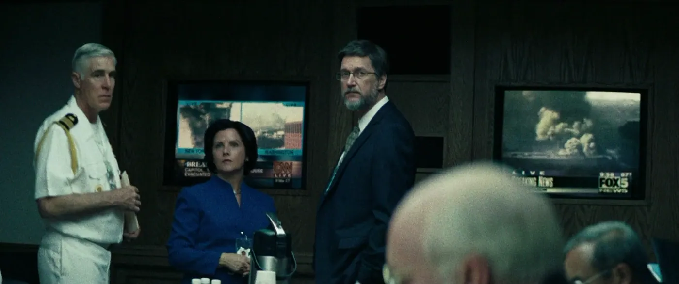 Vice (2018), Adam McKay — plan moyen, intérieur, cinq personnages, clair-obscur, lumière froide : bureau, uniforme blanc, tailleur bleu, téléviseur, thermos, lunettes, costume, cravate