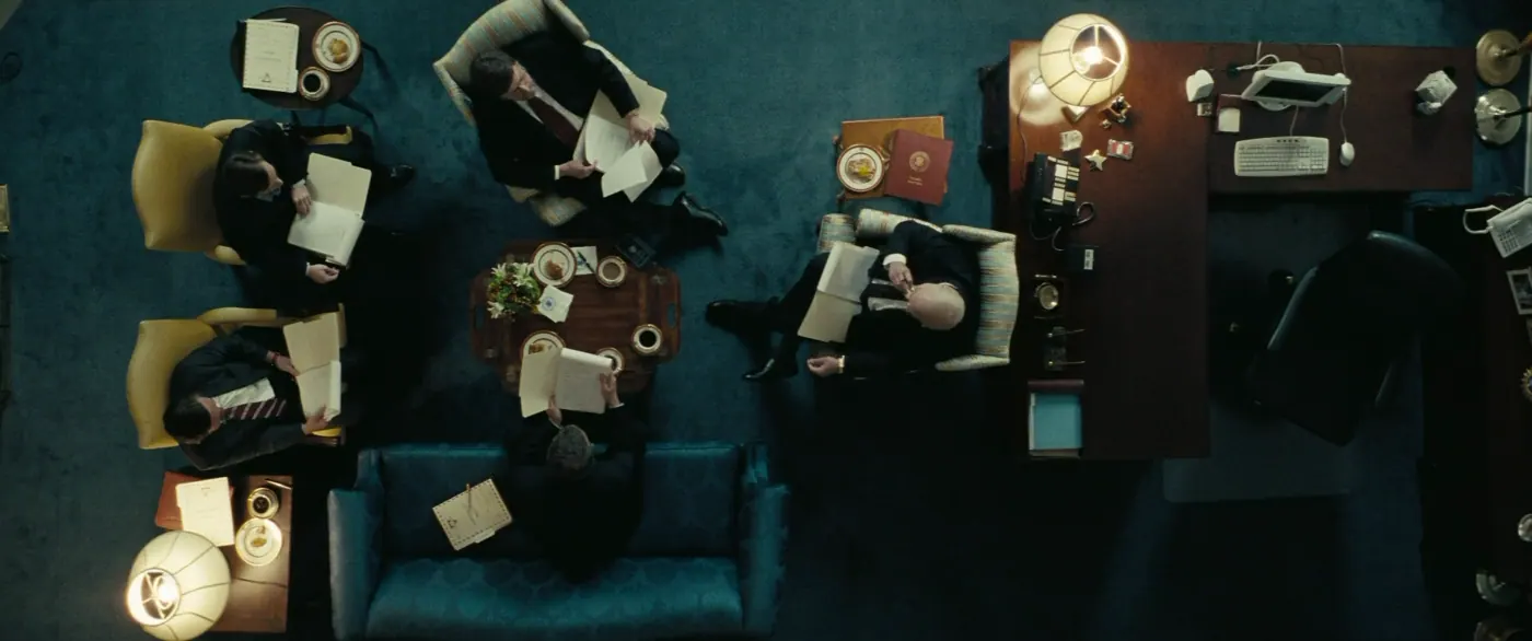 Vice (2018), Adam McKay — plan d’ensemble, intérieur, cinq personnages, lumière chaude, clair-obscur : bureau, fauteuil, canapé, lampe, table basse, costume, cravate, lire