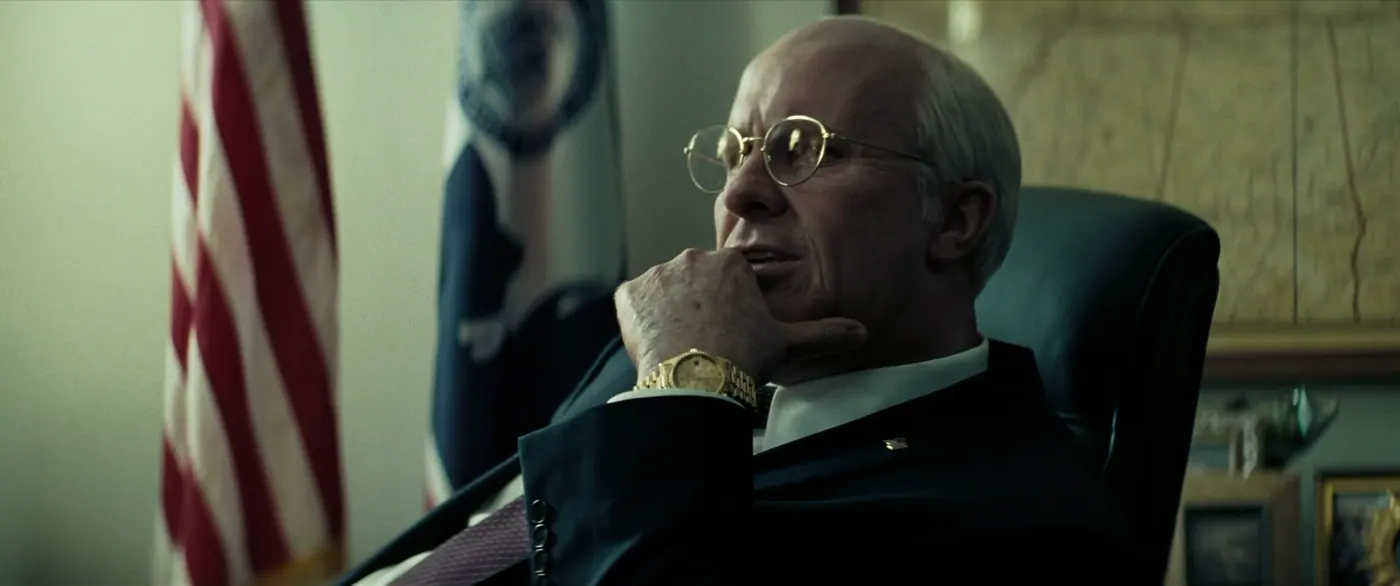 Vice (2018), Adam McKay — plan rapproché, intérieur, un personnage, lumière froide : bureau, drapeau américain, drapeau, montre, lunettes, fauteuil, costume, cravate