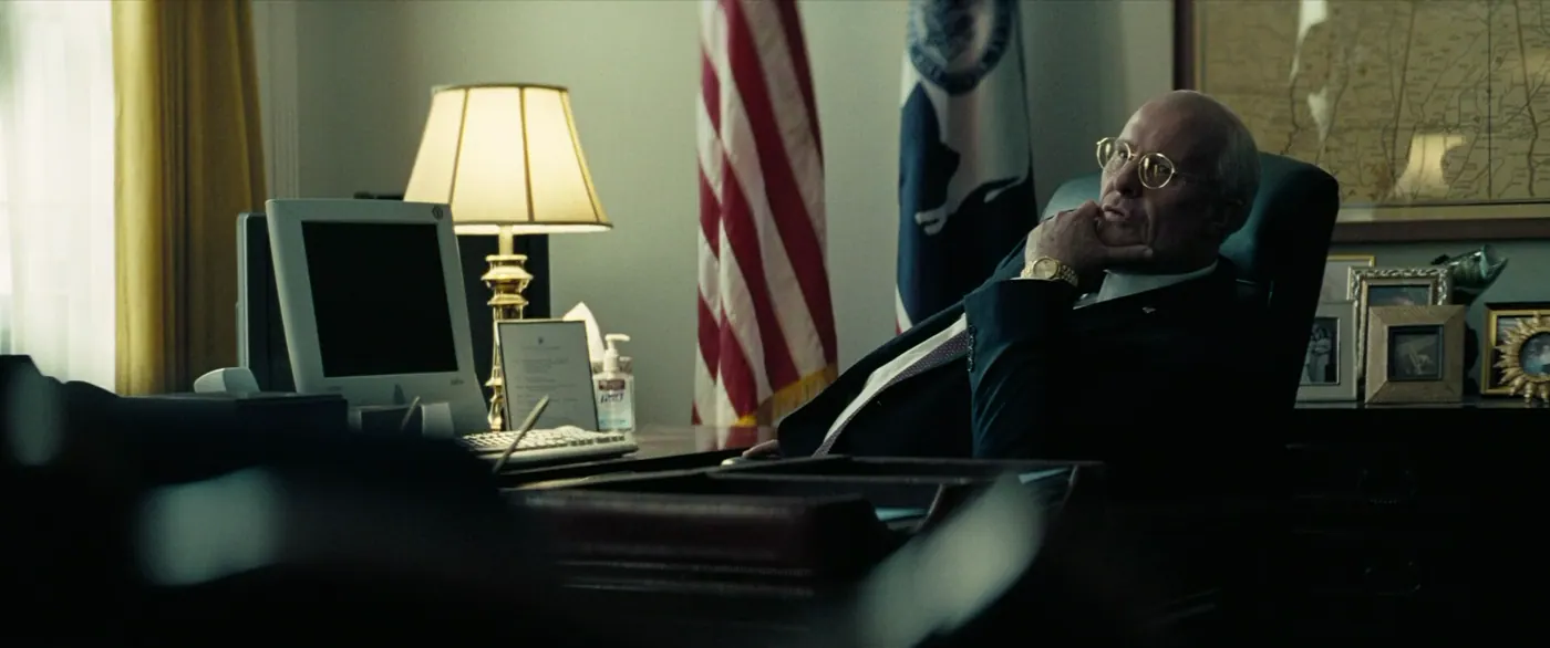 Vice (2018), Adam McKay — plan large, jour, intérieur, un personnage, lumière de fenêtre, lumière chaude : bureau, ordinateur, clavier, lampe, drapeau américain, costume, cravate