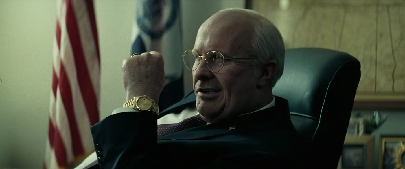 Vice (2018), Adam McKay — plan rapproché, intérieur, un personnage, lumière froide : bureau, drapeau américain, drapeau, montre, bague, lunettes, costume, cravate, lever le poing