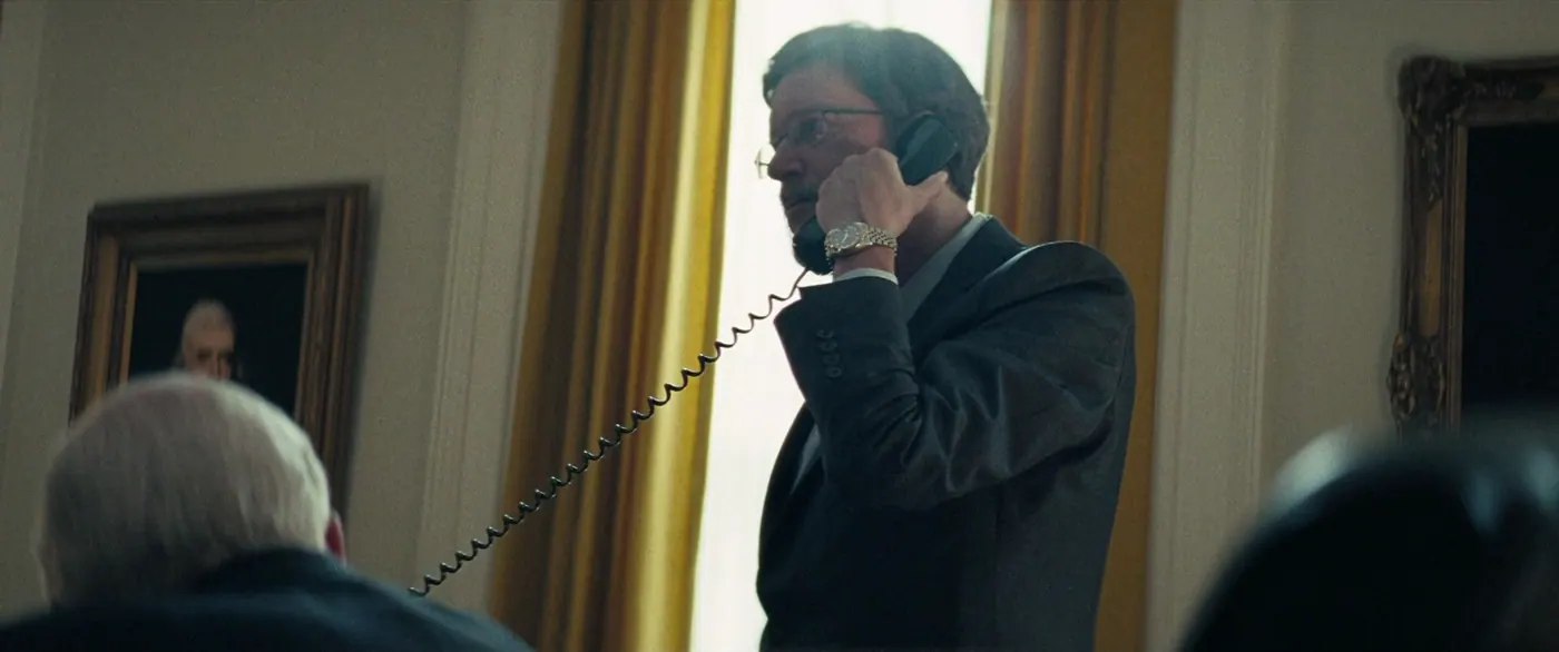 Vice (2018), Adam McKay — plan rapproché, jour, intérieur, trois personnages, contre-jour, lumière de fenêtre : bureau, téléphone fixe, montre, lunettes, rideau, fenêtre, costume, téléphoner